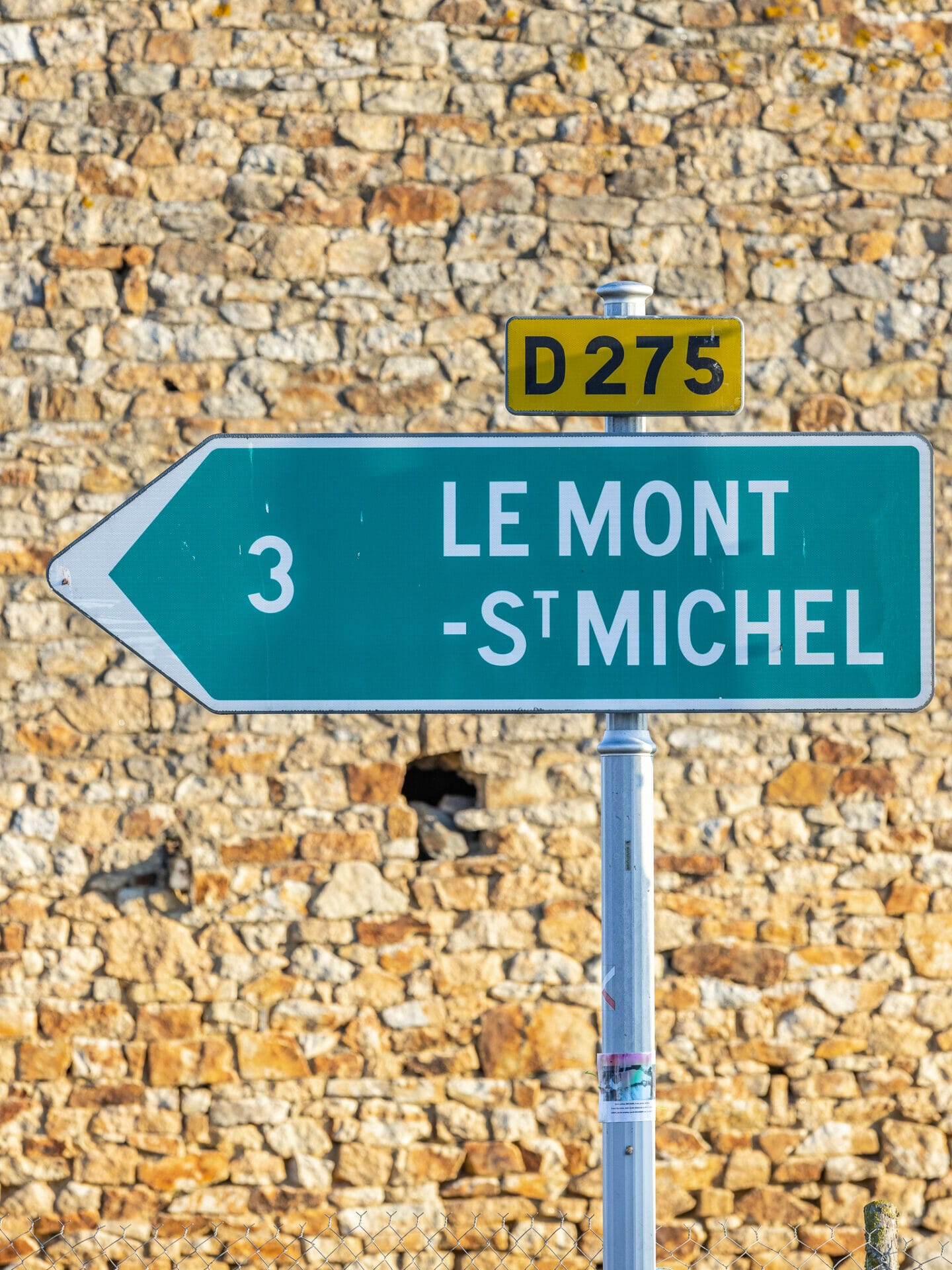 Green street sign pointing left with "Le Mont St Michel" written on it on old stone background